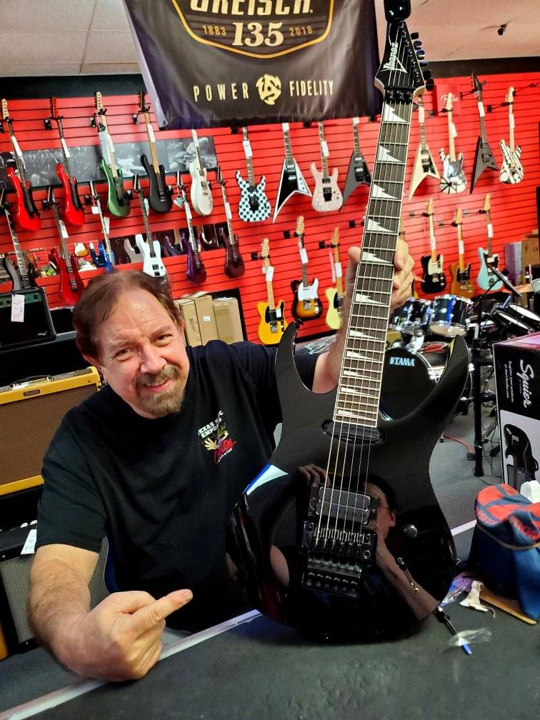 Man in a music shop smiles, pointing at a sleek black electric guitar he's holding. Brightly colored guitars hang on a red wall in the background.