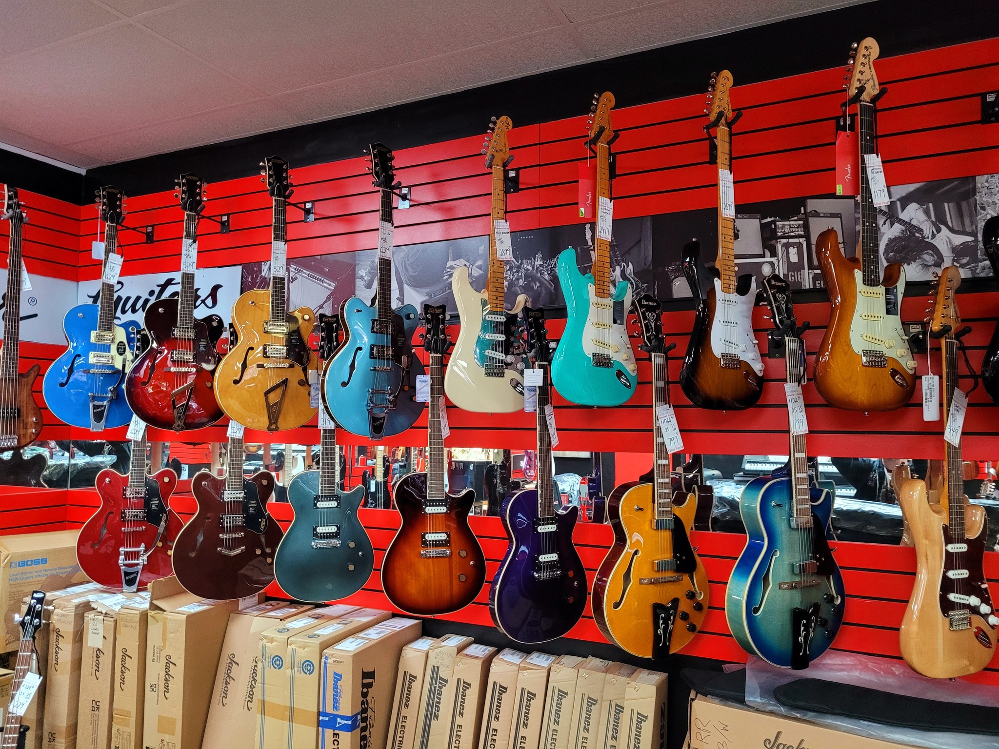 A colorful array of electric guitars hangs on a vibrant red wall in a music store. The variety of colors and styles creates a lively and inviting atmosphere. Boxes are stacked below.