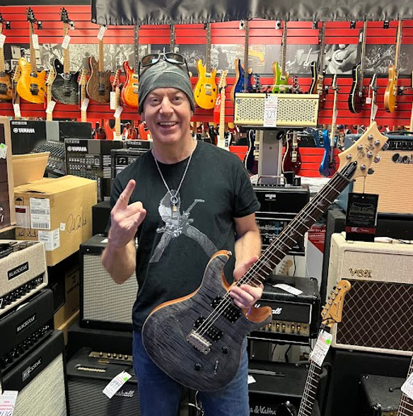 A smiling man in a beanie and black T-shirt holds a dark electric guitar in a music store. Vibrant guitars line the red walls, amplifiers below.